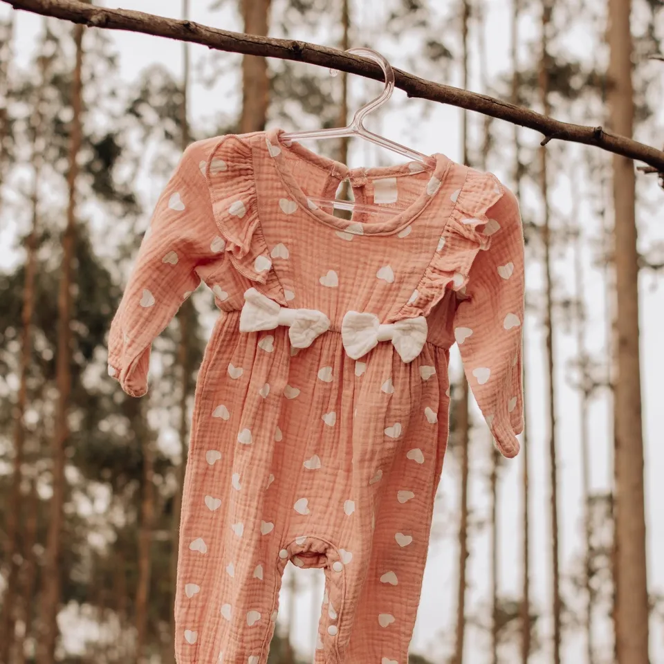 Jumpsuit bayi lengan panjang warna persik bermotif hati putih, dengan ruffle dan dua pita